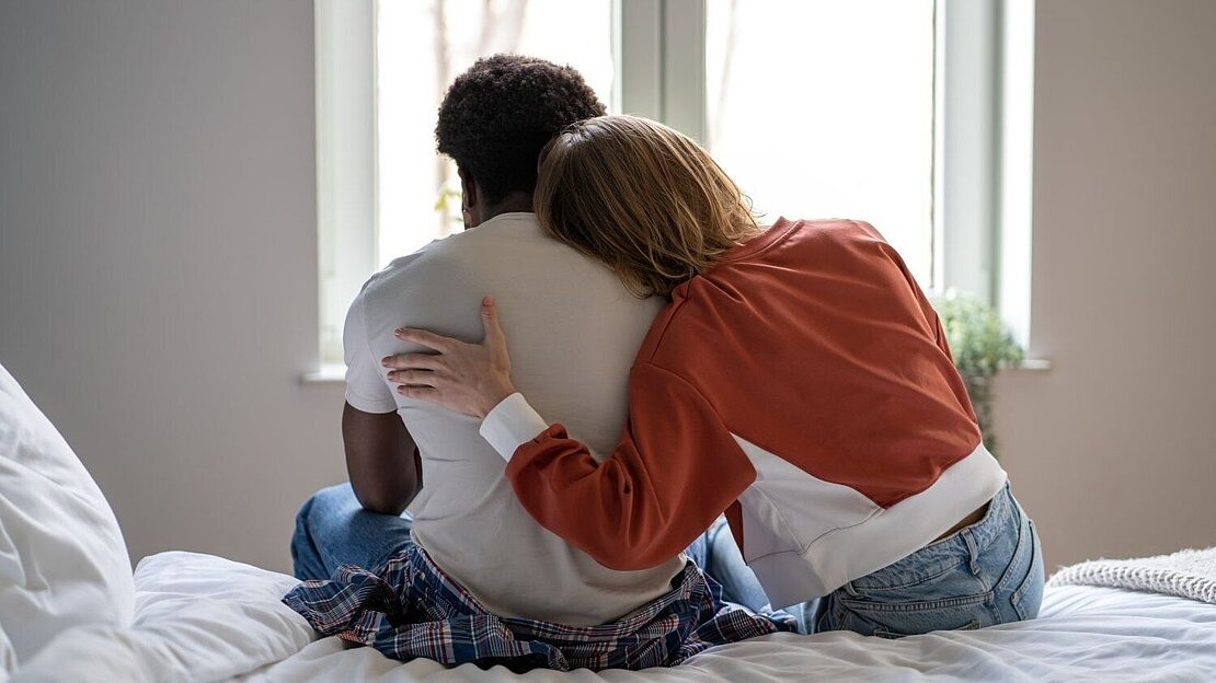 Eine Person legt den Arm um ihren Partner, während beide nebeneinander auf einem Bett sitzen. Symbolbild für emotionale Belastung und Unterstützung in Beziehungen bei Glücksspielsucht.